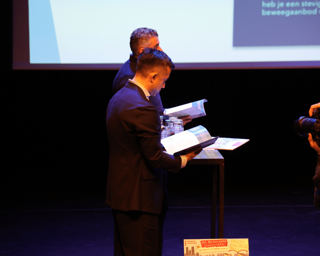 De wethouders Sofyan Mbarki en Alexander Scholtes die de handleiding in ontvangst nemen en doorbladeren
